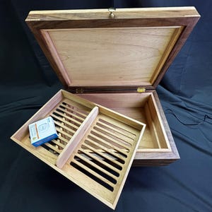 May include: A rectangular wooden humidor with a dark walnut exterior and a light cedar interior. The lid is open, revealing a removable cedar tray with slotted ventilation slats and a central divider. A small blue and white digital hygrometer sits inside.
