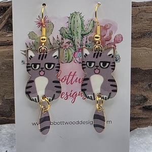 May include: A pair of cat-shaped earrings with gold-colored hooks. The earrings feature a gray cat design with black stripes, white bellies, and a small dangling tail. The earrings are on a white card with the text "Bottwood Designs".