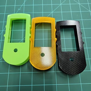 Op de afbeelding: Drie 3D-geprinte kunststof behuizingen in groen, geel en zwart. Elke behuizing heeft een rechthoekige opening en een klein rond gat.