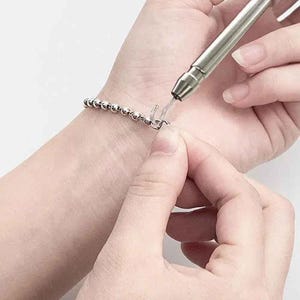 May include: A silver beaded bracelet is being clasped with a jewelry tool. The tool is silver and has a hook-like end. The bracelet is made of small, round, silver beads. The clasp is being closed on a wrist.