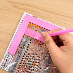 May include: A pink diamond painting tool kit with a pink frame and a pink pen. The kit is being used on a partially completed diamond painting project. The project has a grid of numbers and letters.