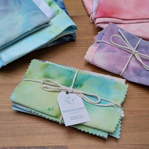 May include: A selection of hand-dyed fabric squares in various colours, including blue, green, pink, and purple. One bundle is tied with twine and a tag that reads "NEEDLELIGHT STUDIO". The fabric has a watercolour effect.