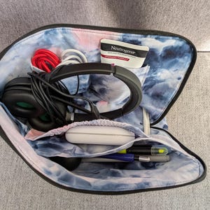 May include: Open black zippered case with a tie-dye blue and white interior. The case contains black headphones, white and red cords, a tube of Neutrogena, a white charging case, and pens. The case is open, revealing the contents.