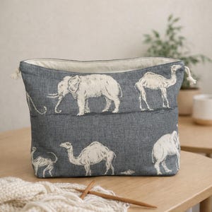 May include: A grey fabric storage basket with a drawstring closure. The basket features a repeating pattern of white elephants and camels. The interior lining is a light cream colour. The basket is on a wooden table.