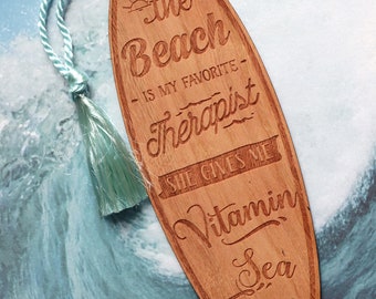 May include: Wooden surfboard bookmark with a blue tassel. The bookmark has the text "The Beach is my favorite therapist. She gives me Vitamin Sea" with a sun and palm tree graphic.