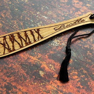 May include: Wooden bookmark shaped like a baseball bat with barbed wire design and the word "Lucille" engraved on the handle. The bookmark has a black tassel.