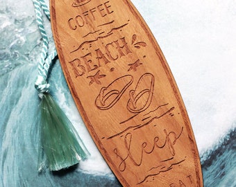 May include: Wooden surfboard-shaped bookmark with the text "Coffee, Beach, Sleep, Repeat" engraved on it. The bookmark has a light brown color and a green tassel.