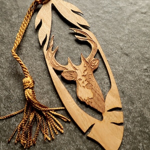 May include: A wooden bookmark shaped like a feather with a deer head carved into it. The bookmark has a gold tassel attached to it.