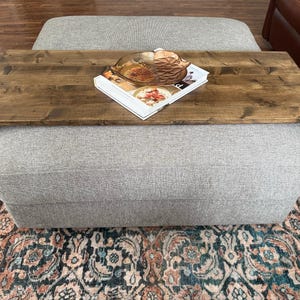 May include: A grey upholstered ottoman with a wooden tray attached to the top. The tray is stained a dark brown and has a book and a decorative bowl on top.