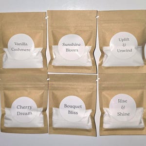 Puede incluir: Seis bolsas de papel kraft marrón que contienen polvo blanco, cada una con una etiqueta redonda blanca. Las etiquetas dicen: Vanilla Cashmere, Sunshine Bloom, Uplift & Unwind, Cherry Dream, Bouquet Bliss y Rise & Shine. Dos filas.