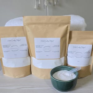 May include: Three brown paper bags of natural laundry detergent with white labels, and a small green bowl with a scoop. The bags are filled with white powder. The text on the label reads "Natural Laundry Detergent".