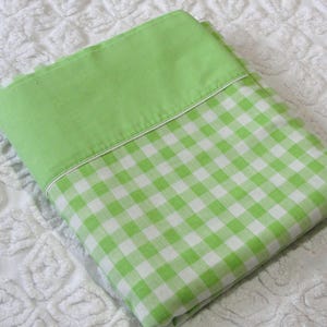 May include: A folded green and white gingham patterned pillowcase with a solid green top section. The pillowcase is on a white textured surface.