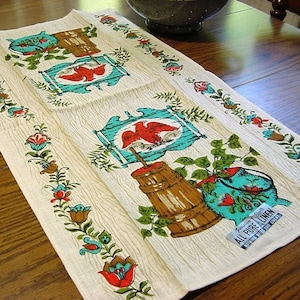 May include: Cream-colored linen towel with a repeating pattern of red birds, turquoise frames, and brown barrels. Floral border in red, blue, and brown. The towel is labeled "All Pure Linen".
