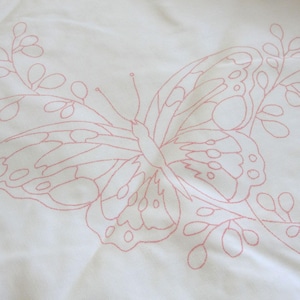 May include: A white fabric surface featuring a delicate, light pink line art embroidery pattern of a large butterfly centered among leafy branches. The design consists of thin, continuous outlines that define the wings and foliage.