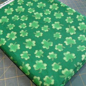 May include: A folded piece of green fabric with a repeating pattern of light green shamrocks. The shamrocks have a glittery texture. The fabric is likely for crafting or sewing projects, perfect for St. Patrick's Day decorations or apparel.