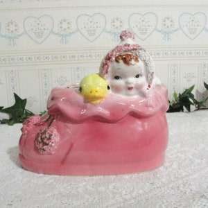 May include: A vintage ceramic planter in the shape of a baby's shoe, painted in a vibrant pink. The planter features a smiling baby face and a yellow duck. Decorative floral accents adorn the shoe's exterior, creating a charming, nostalgic aesthetic.