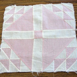 May include: A square quilt block featuring a central pink and white gingham pattern, surrounded by white fabric and geometric shapes. The block is made of stitched fabric pieces, with a handmade, vintage aesthetic.