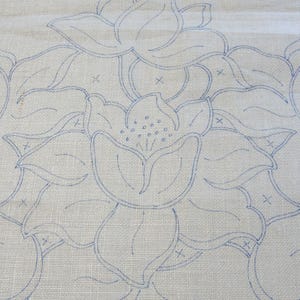 May include: Close-up of a cream textile with a repeating floral pattern. The design features large, stylized flowers outlined in light blue thread. The fabric has a subtle, textured appearance.
