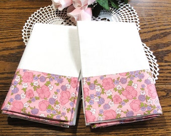 May include: Two white hand towels with a pink floral border featuring pink and purple flowers and green leaves. The towels are folded and stacked, showcasing the patterned trim. The background includes a white lace doily and wooden surface.