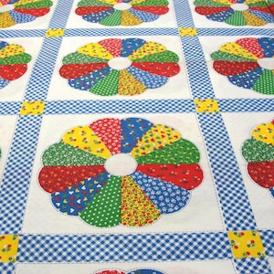 May include: A colorful quilt featuring a repeating floral pattern. Each flower is composed of various fabric pieces in red, yellow, blue, and green. The quilt has a white background with blue gingham and yellow square accents.