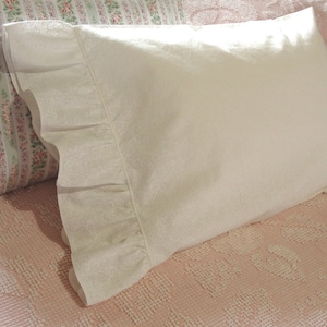 May include: A white pillow with a ruffled edge rests on a pink patterned bedspread. Another pillow with a floral pattern is visible in the background. The pillow has a subtle, textured pattern.