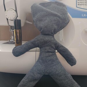 May include: A gray heathered fabric doll with a rounded head and outstretched limbs, positioned next to a small amber glass roller bottle with a black cap, set against a white sewing machine with visible dials and text.