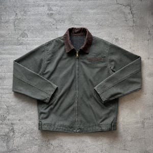 Könnte beinhalten: Dunkelgrüne Jacke mit braunem Kragen und Reißverschluss. Die Jacke hat eine braune Brusttasche und zwei Seitentaschen. Sie besteht aus strapazierfähigem Material und hat ein klassisches Design.