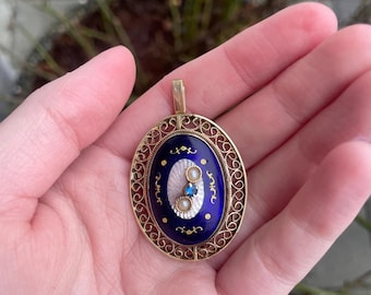 May include: An oval pendant with a gold filigree frame. The pendant features a deep blue enamel center with a white oval design, adorned with a pearl, a blue gemstone, and gold accents. The pendant is suspended from a gold bail.