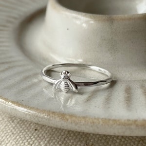 May include: A silver ring with a bee design. The ring is on a white ceramic dish.