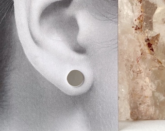 May include: A close-up of a silver stud earring worn in a person's ear. The earring is a simple, round shape and is set against a backdrop of a white card with the text "annie lespérance" printed on it.