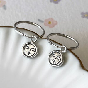 May include: A pair of silver hoop earrings with a small silver charm hanging from each hoop. The charms are round and feature a face with closed eyes.