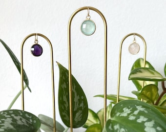 Puede incluir: Tres soportes de metal dorado con tapas redondas, cada uno con un colgante de piedra preciosa de color diferente. Los colgantes son de amatista, aguamarina y cuarzo rosa. Los soportes están delante de una planta en maceta con hojas verdes y blancas.