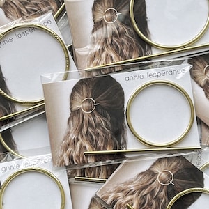 May include: Gold hair clips and a gold bar hair stick. The hair clips are round and have a hammered texture. The hair stick is straight and smooth. The hair clips and stick are displayed on white cards with the text "annie lesp&eacute;rance" printed on them.
