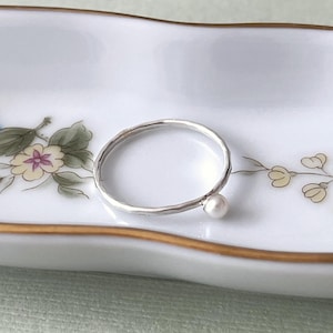 Peut inclure: Une délicate bague en argent avec une petite perle blanche en accent. La bague est sur un plat en porcelaine blanche avec des motifs floraux.