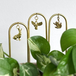 May include: Three gold metal plant stakes with different animal charms. The charms are a rabbit, an owl, and a frog. The stakes are on a white background with a green plant in the foreground.