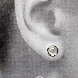 May include: A close-up of a person's ear wearing a silver earring with a white pearl. The earring is a simple design with a small circle around the pearl.