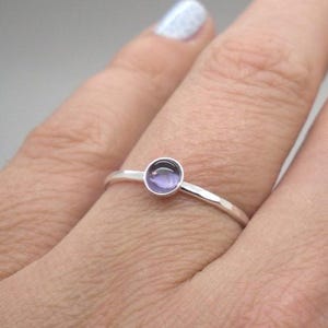 May include: A silver ring with a small, round, purple gemstone. The ring is simple and elegant, with a thin band and a bezel setting for the gemstone. The ring is shown on a person's finger.