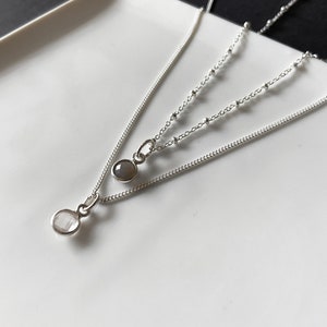 May include: Two silver necklaces with small oval-shaped stones. The top necklace has a gray stone and the bottom necklace has a white stone.