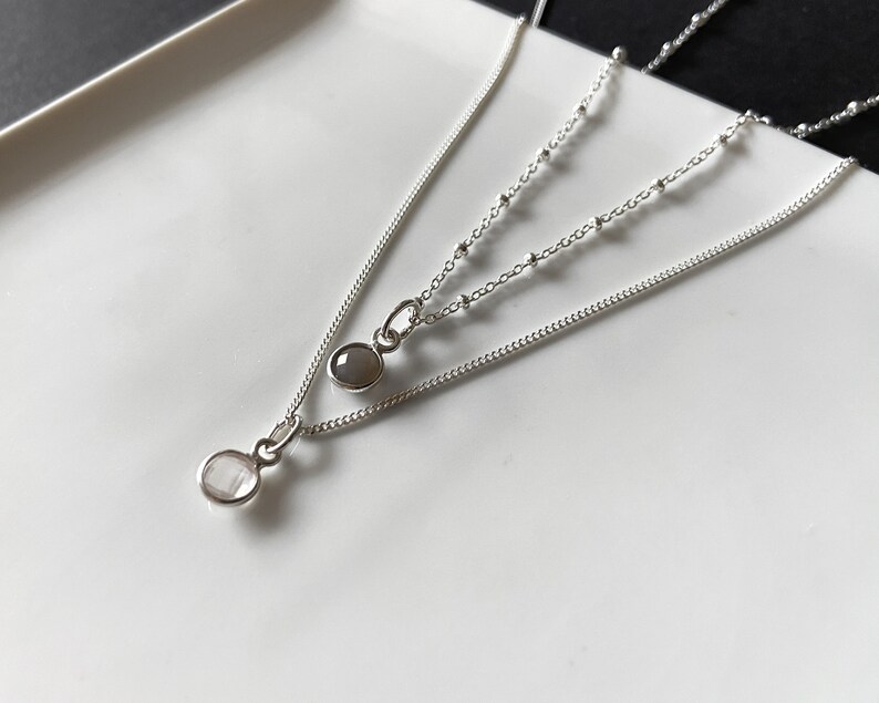 May include: Two silver necklaces with small oval-shaped stones. The top necklace has a gray stone and the bottom necklace has a white stone.