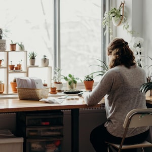 May include: A person sits at a desk in front of a window, working on a project. The desk is cluttered with plants, a basket of papers, and other supplies. The person is wearing a grey sweater and has their back to the camera.