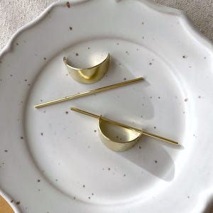 Pu&ograve; includere: Due fermagli per capelli dorati, uno &egrave; un semicerchio e l'altro &egrave; una barra dritta. I fermagli sono su un piatto bianco.