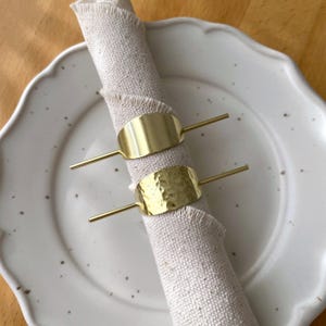 Pu&ograve; includere: Due anelli per tovaglioli in oro, uno con finitura liscia e l'altro con una texture martellata, sono posizionati su un tovagliolo di lino bianco.