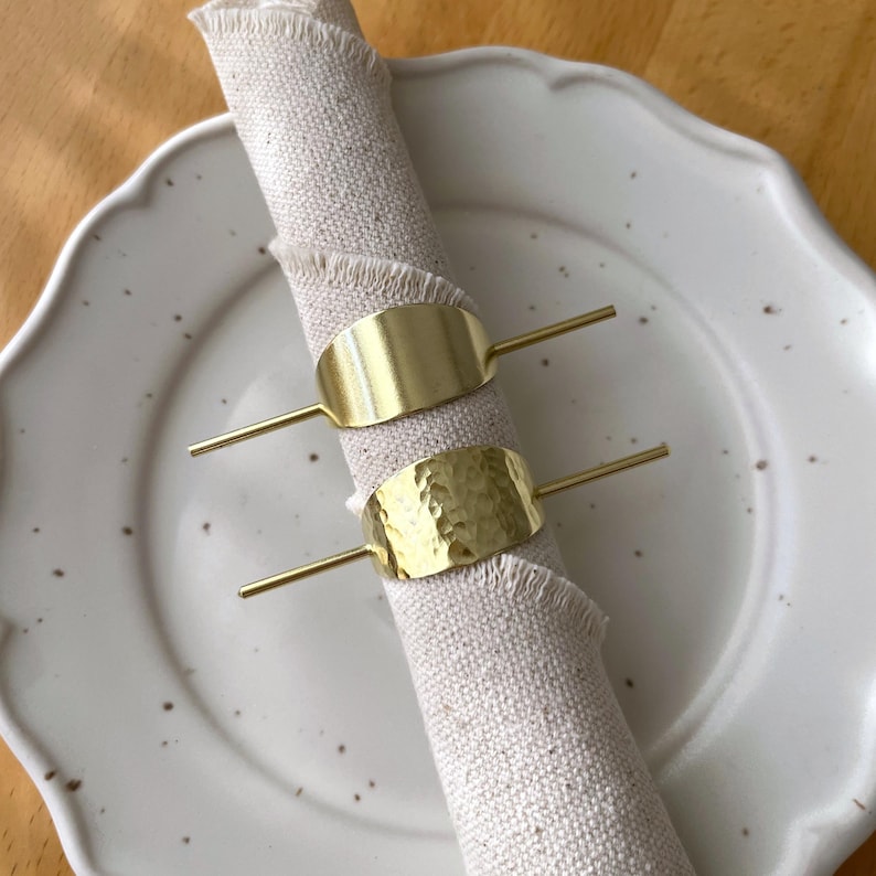 Pu&ograve; includere: Due anelli per tovaglioli in oro, uno con finitura liscia e l'altro con una texture martellata, sono posizionati su un tovagliolo di lino bianco.