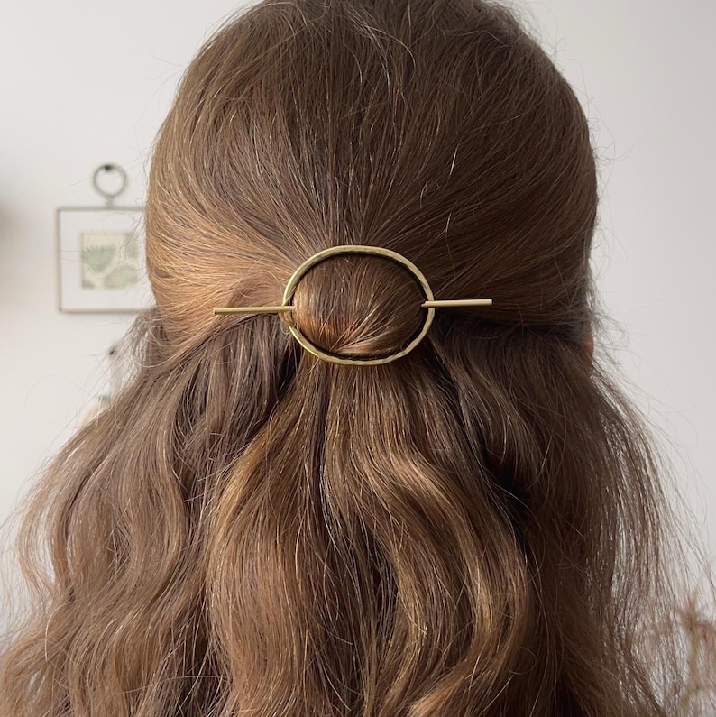 May include: A gold oval hair clip with a straight bar across the center. The clip is in a woman's brown hair.