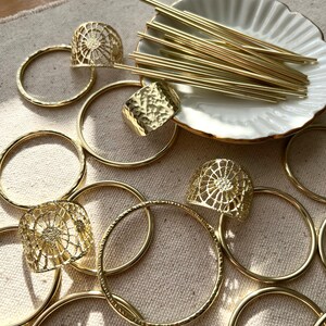 May include: A collection of gold-toned metal rings, bangles, and decorative pieces. The items are arranged on a beige fabric surface. Some of the pieces have a textured finish, while others have a delicate, openwork design.