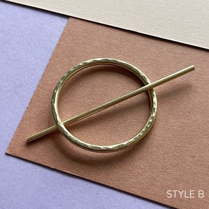 May include: Gold metal hair clip with a hammered circle and a straight bar. The clip is on a brown and purple background. STYLE B.
