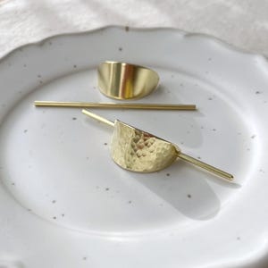 Pu&ograve; includere: Due fermagli per capelli dorati, uno liscio e uno con una texture martellata, sono esposti su un piatto bianco. I fermagli sono progettati per tenere i capelli in posizione.