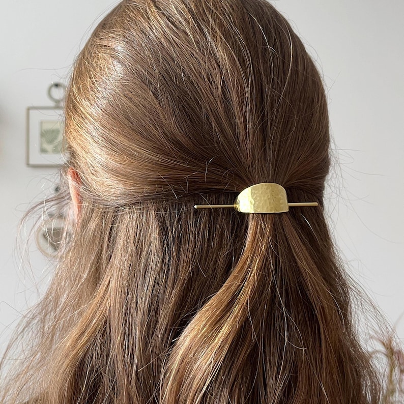 Pu&ograve; includere: Un fermaglio per capelli dorato con una texture martellata. Il fermaglio ha la forma di un semicerchio ed &egrave; attaccato a una sottile barra dritta.