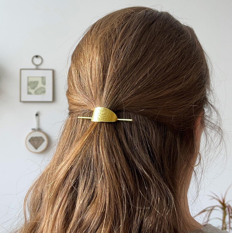 Pu&ograve; includere: Un fermaglio per capelli dorato con una texture martellata e una forma curva. Il fermaglio &egrave; nei capelli di una persona con capelli lunghi e castani.