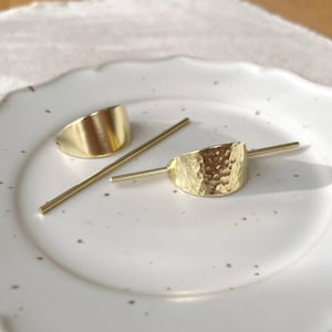 Pu&ograve; includere: Due fermagli per capelli dorati, uno con un design curvo liscio e l'altro con una texture martellata. Entrambi i fermagli sono abbinati a un bastoncino per capelli in metallo dorato.